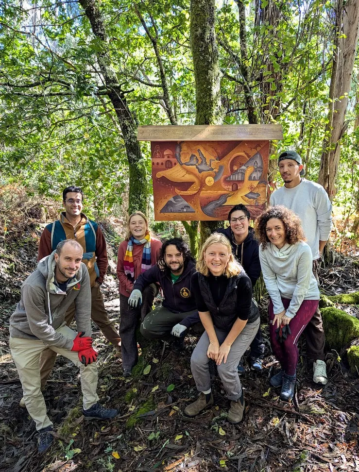 Eight volunteers in work gloves posing in the forest around a painted panel mounted under a small wooden roof on a tree