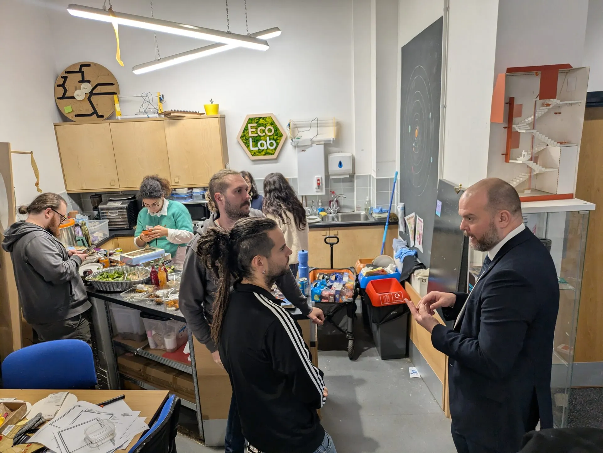 Un hombre en traje conversa con visitantes en la cocina del makerspace Eco Lab, con comida servida en un carro