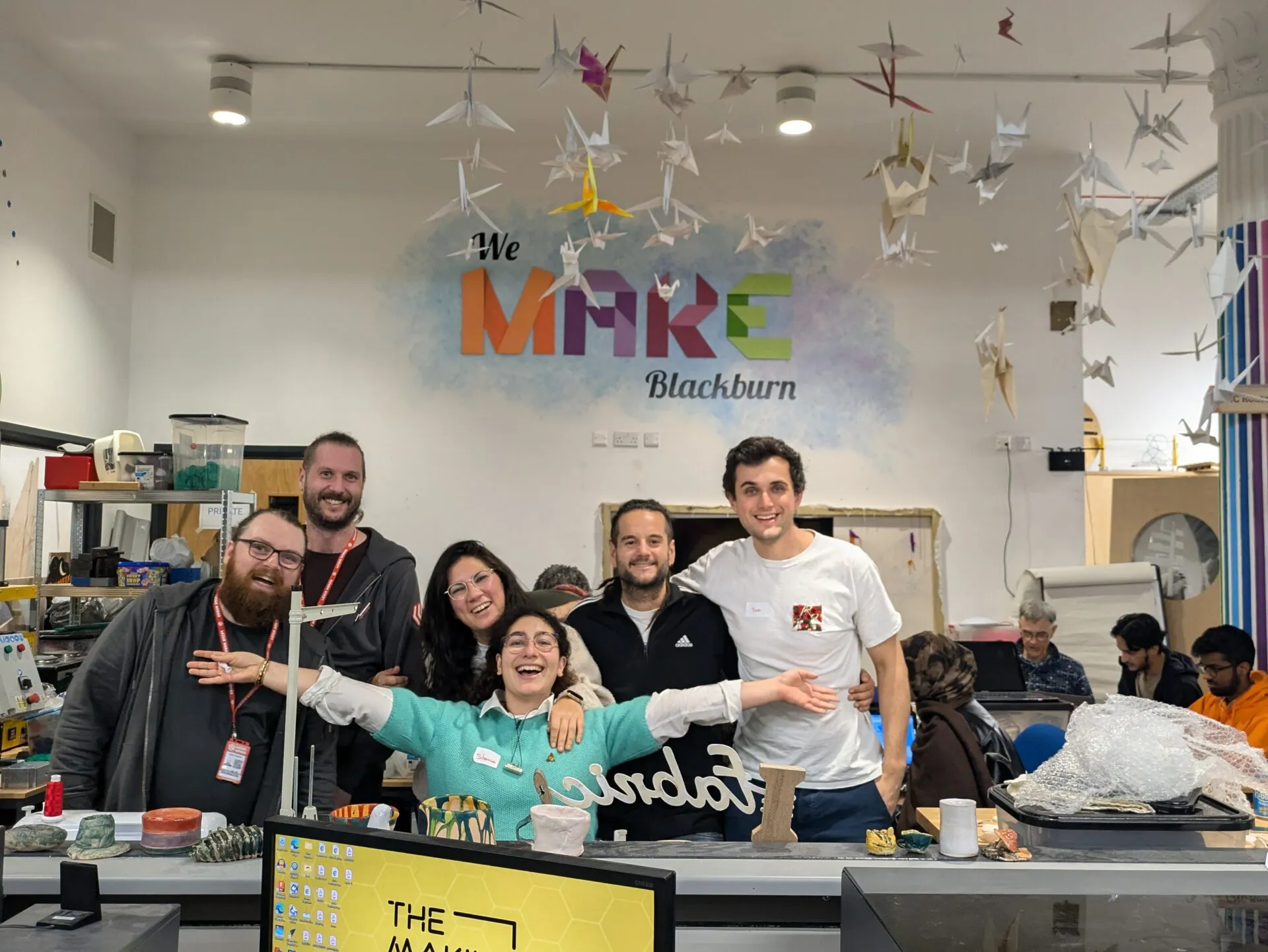 Seis personas riendo juntas en un makerspace bajo un logo en la pared que dice ‘We Make Blackburn’, con grullas de papel colgando del techo