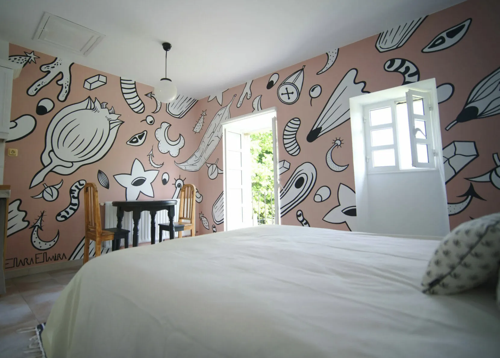 Un dormitorio con una gran cama blanca y paredes pintadas con dibujos en blanco y negro sobre fondo rosa, con puertas de balcón abiertas