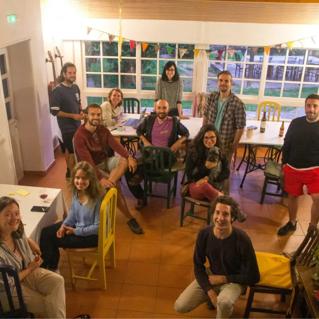 Unha ducia de persoas en cadeiras desparelladas nunha sala luminosa con bandeiroliñas, mirando cara á cámara