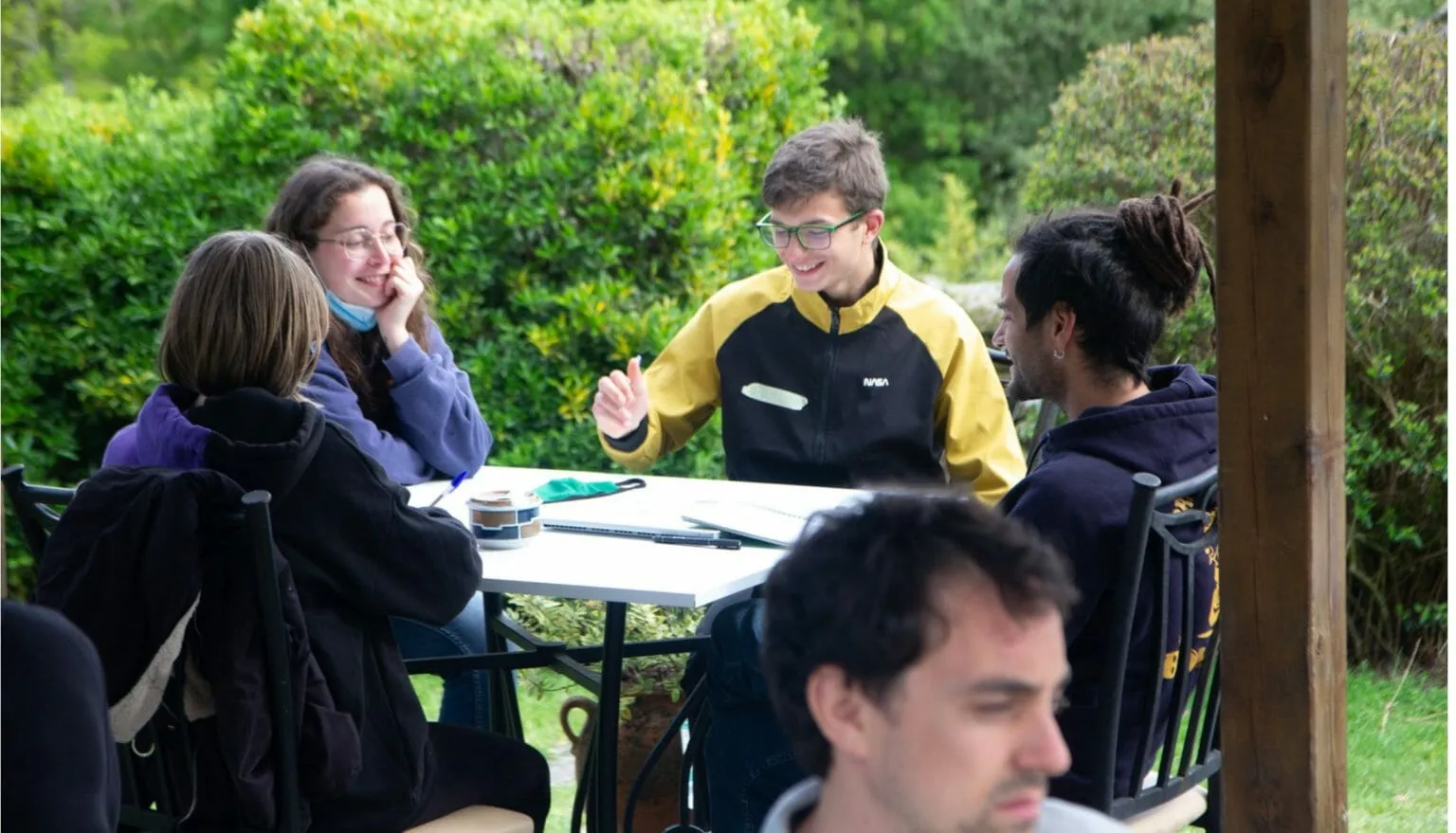 Jóvenes conversando alrededor de una mesa blanca al aire libre, con cuadernos y bolígrafos frente a un seto verde