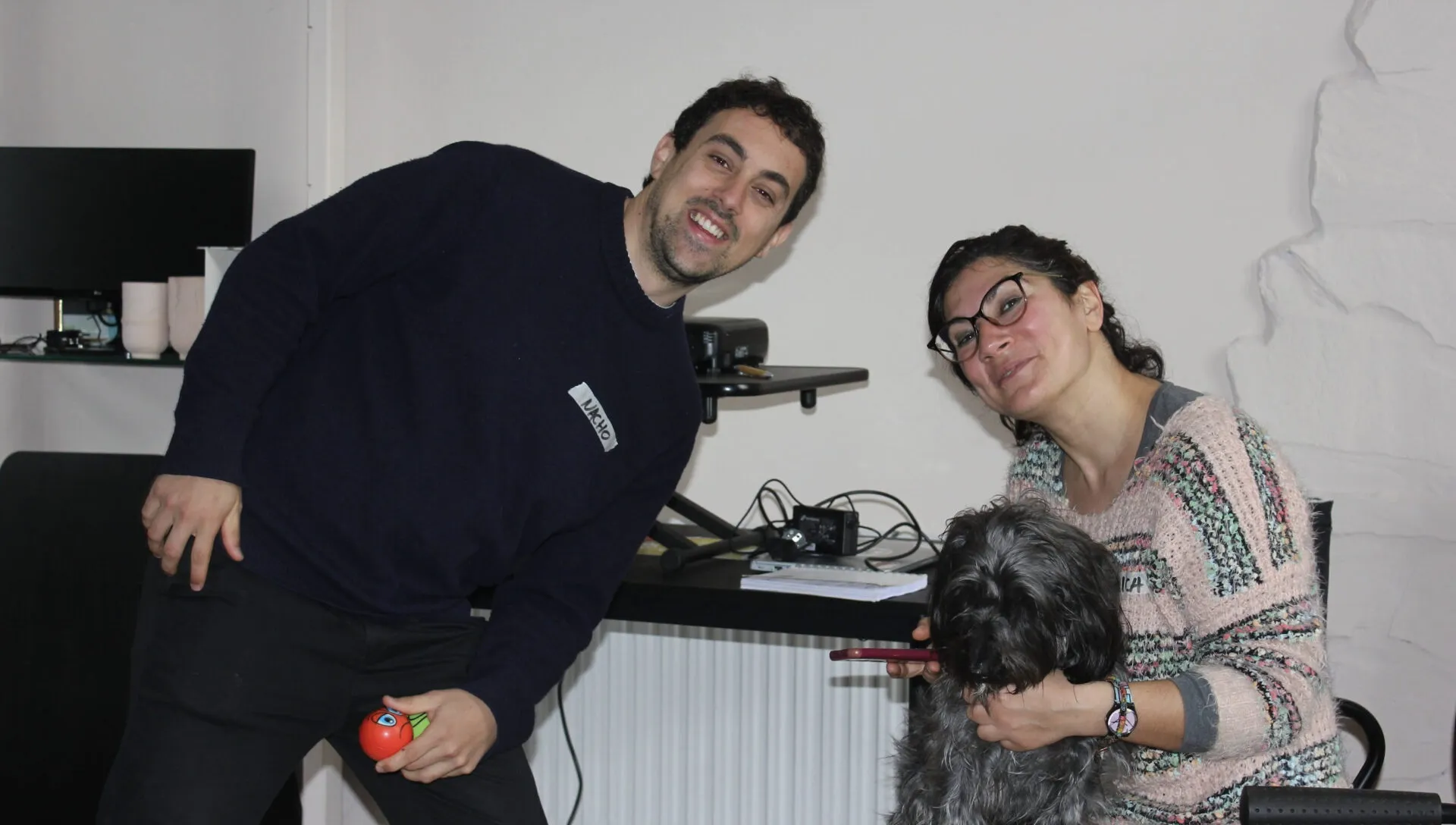 Un hombre sonriente con jersey azul marino inclinado junto a una mujer con gafas que abraza a un perro gris peludo en una oficina