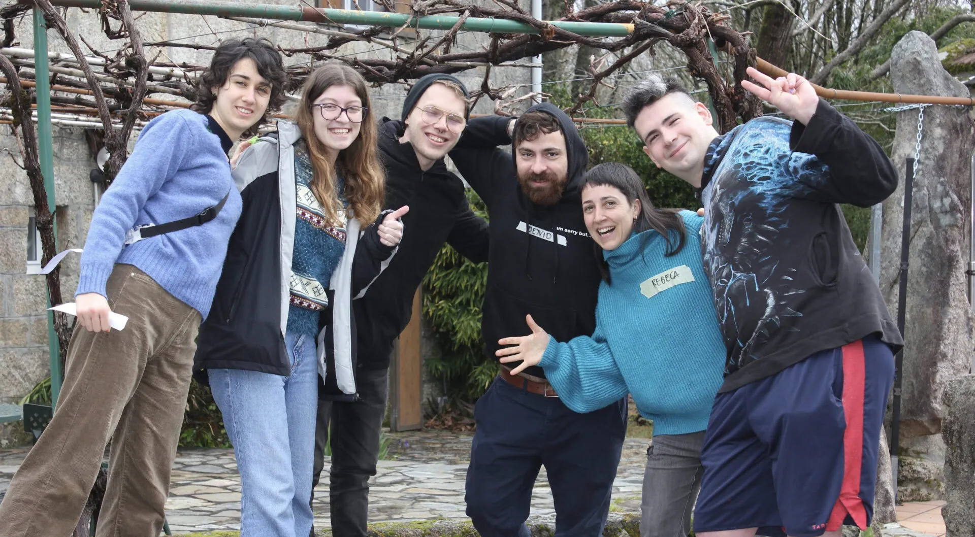 Seis jóvenes posando en broma, abrazados bajo una parra sin hojas; una lleva una etiqueta de papel con el nombre Rebeca
