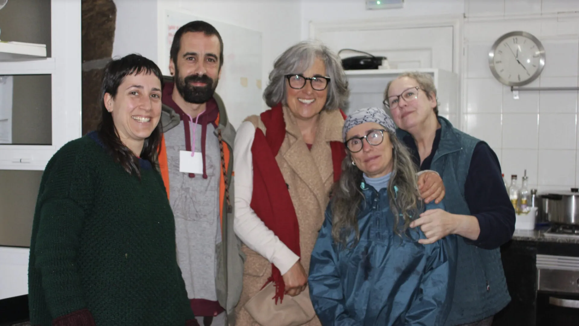 Cinco personas sonriendo abrazadas en una cocina comunitaria de azulejos blancos con un reloj de pared al fondo