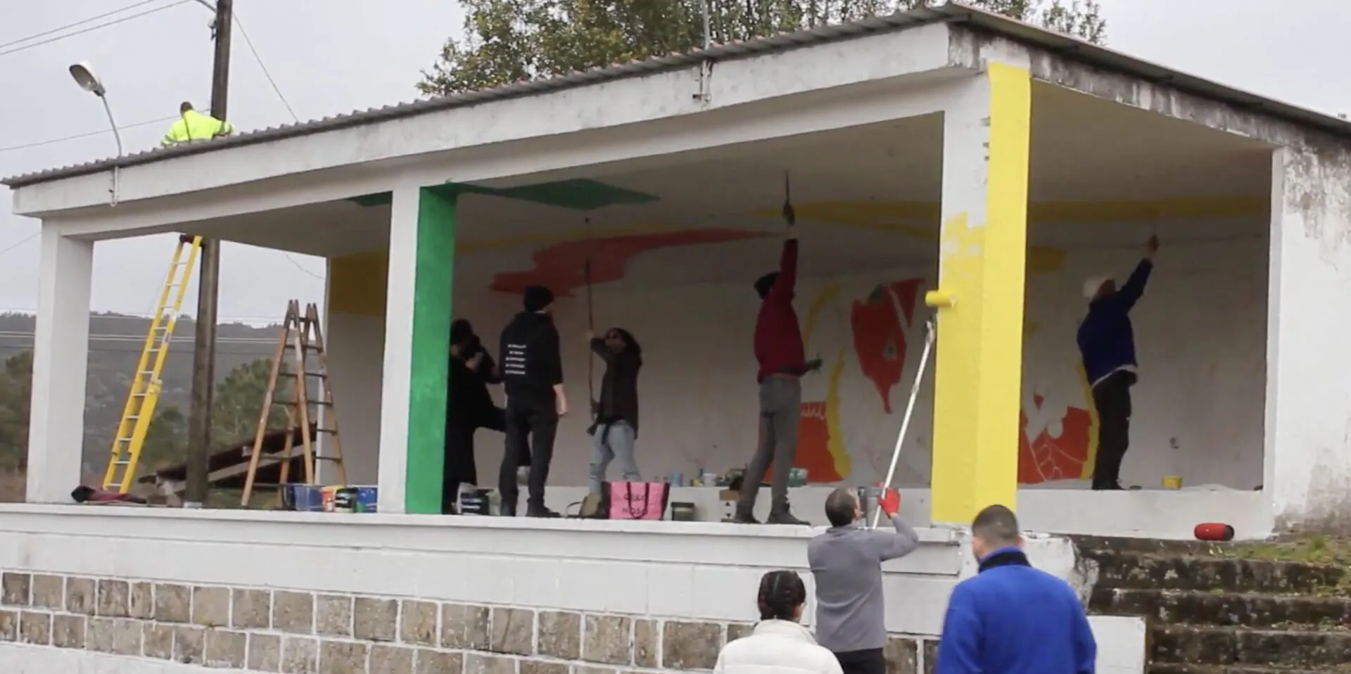 Seis personas pintando un gran mural de colores en el palco de la aldea, con escaleras y botes de pintura alrededor