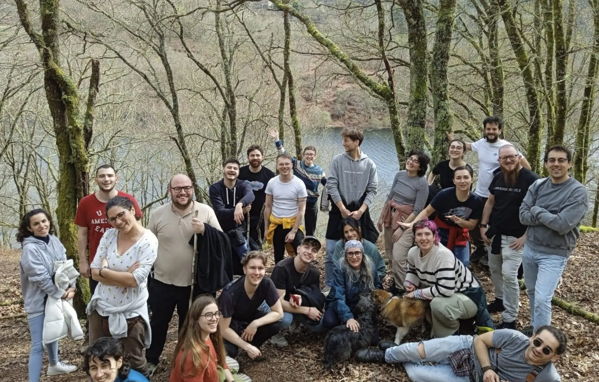 Unas veinticinco personas jóvenes posando entre árboles desnudos y cubiertos de musgo en una ladera sobre un embalse