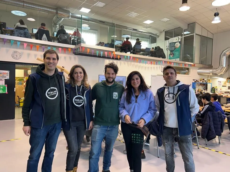 Cinco persoas posando nun fab lab, tres con sudadeiras de FABLAB León, con bandeiriñas e un altillo de mesas con ordenadores detrás