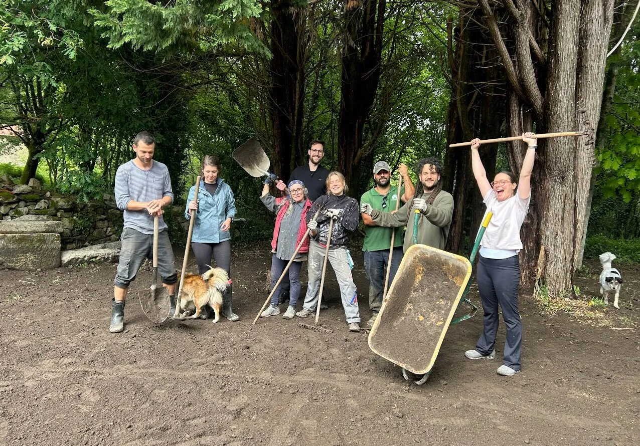 Ocho personas posando con palas, rastrillos y una carretilla en alto sobre tierra recién removida, con dos perros al lado