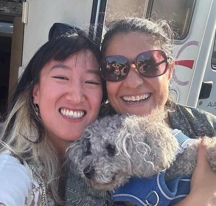 Selfi de dos mujeres mejilla con mejilla sosteniendo un perrito gris de pelo rizado delante de una autocaravana