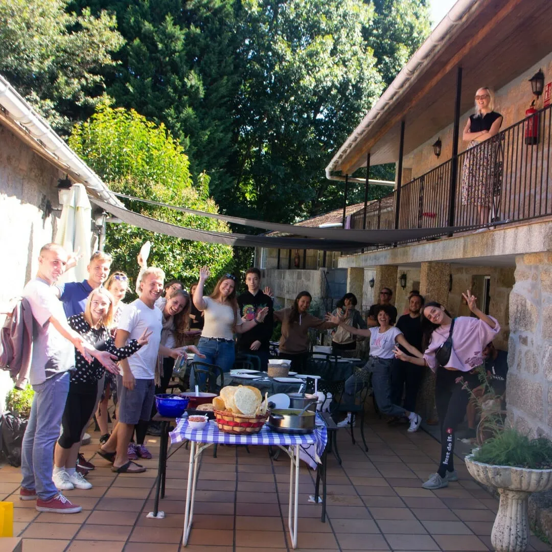 Unas quince personas saludando y posando en la terraza de una casa de piedra junto a una mesa con pan y comida compartida