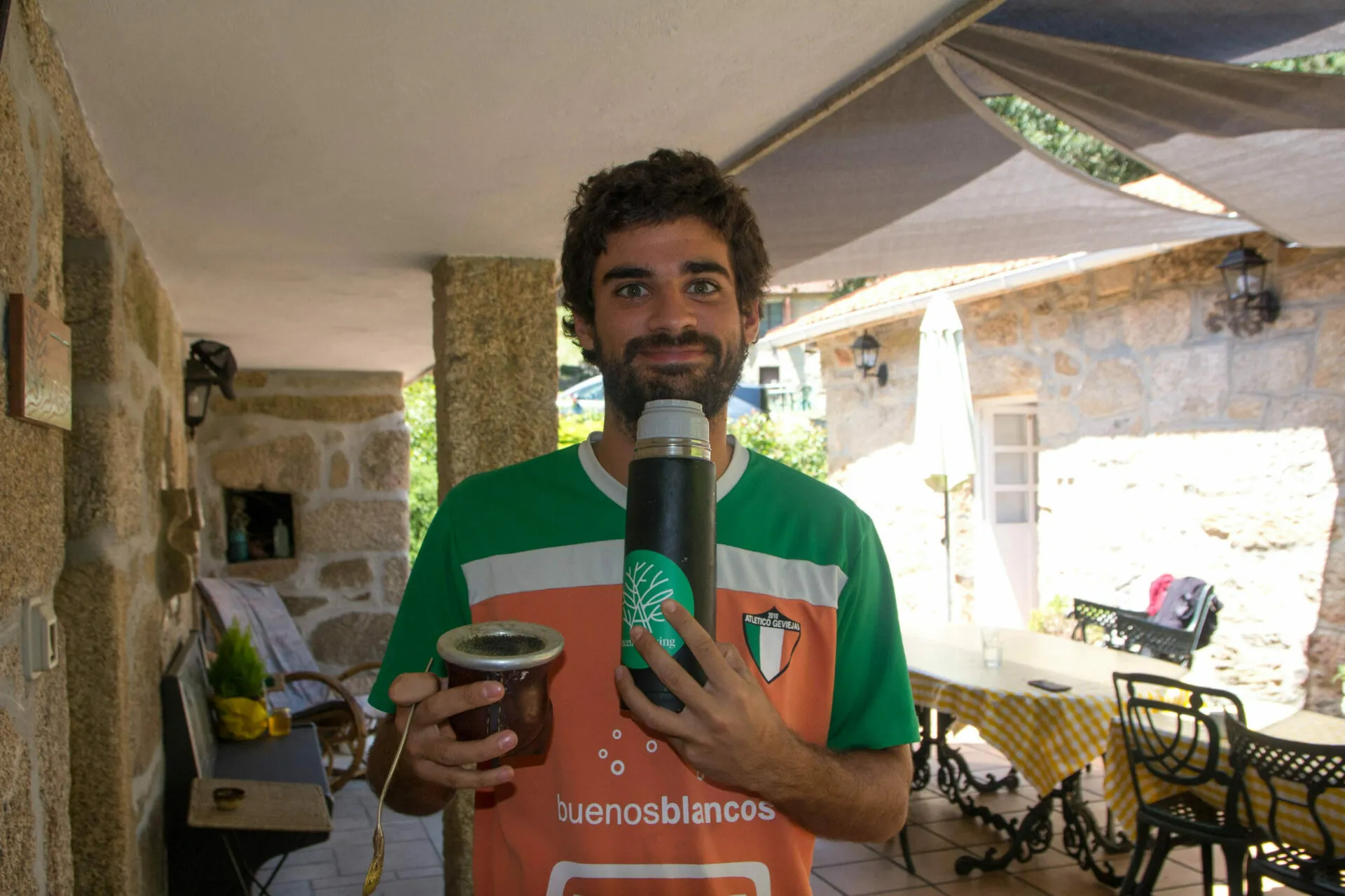 Mozo barbudo cunha camiseta de fútbol laranxa e verde cun mate e un termo nunha terraza de pedra