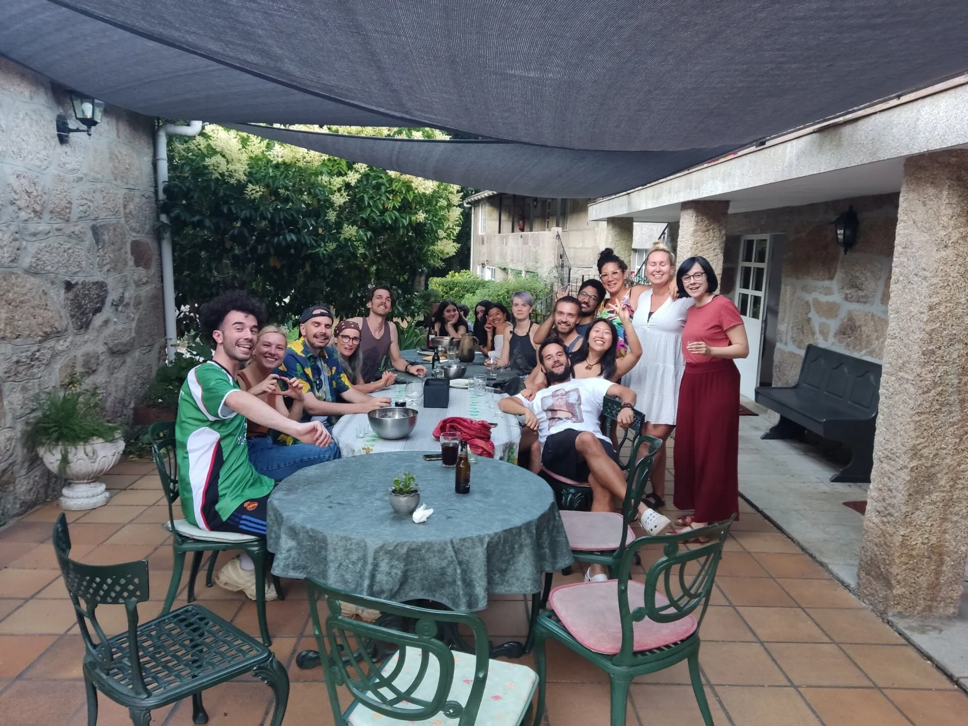 Una mesa larga de amigos en una terraza de piedra cubierta, algunos sonriendo a la cámara, con un mantel verde en la mesa delantera
