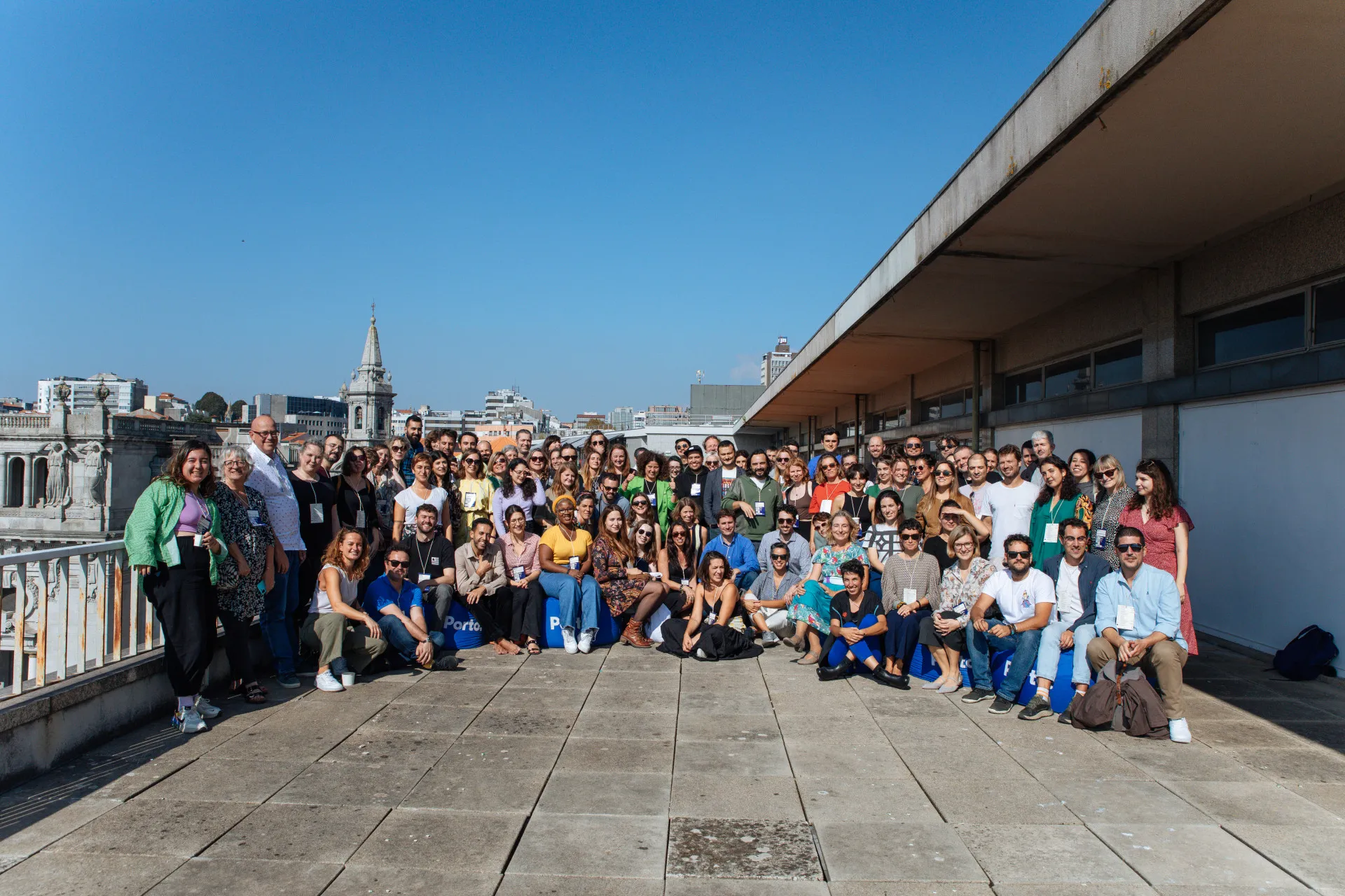 Unha multitude de máis de cen persoas posando nunha terraza de azotea pavimentada baixo un ceo azul, con edificios de cidade detrás