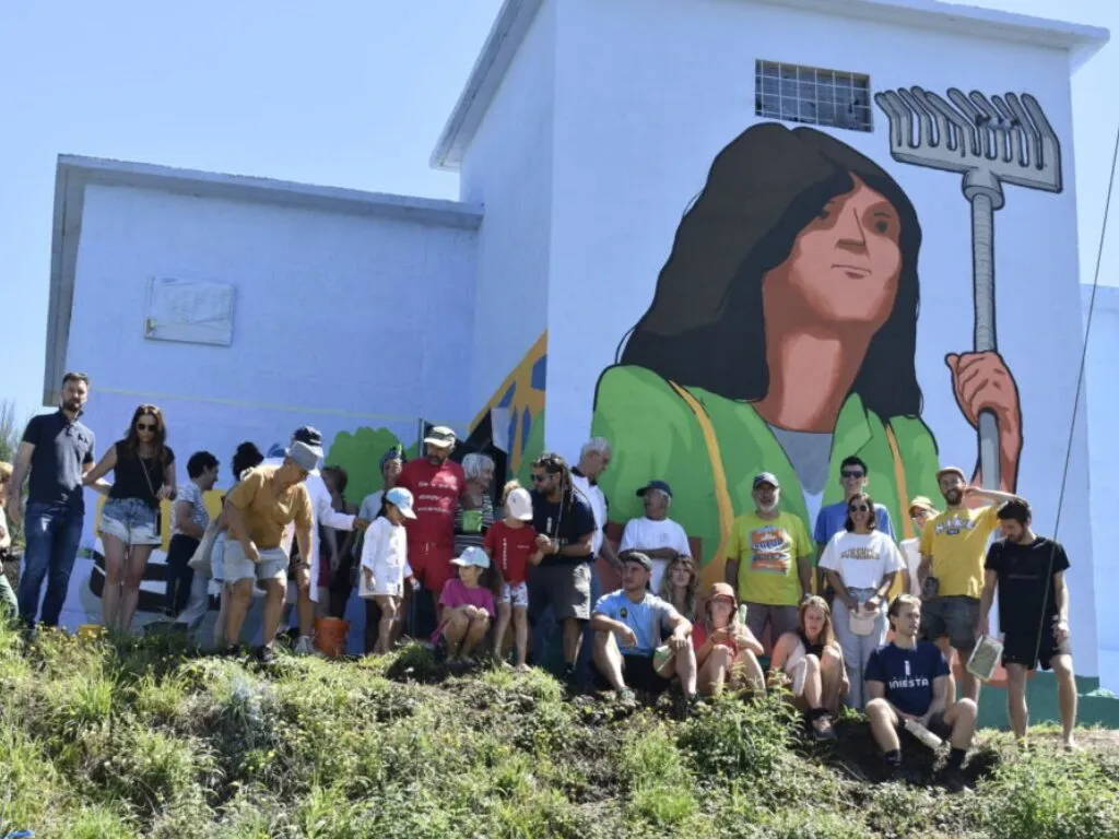 Veciñanza e colivers reunidos baixo un mural dunha muller cun angazo pintado nun edificio branco