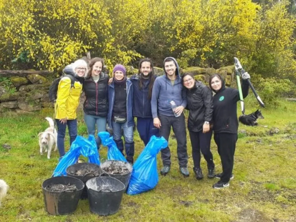 Sete persoas con chuvasqueiros tras caldeiros e bolsas azuis de residuos recollidos, con dous cans e un arbusto de flores amarelas
