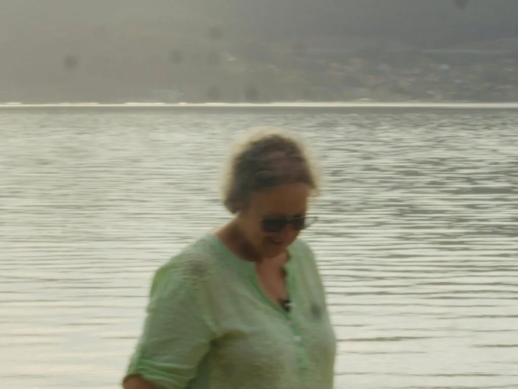 Imaxe desenfocada dunha muller con blusa verde clara e lentes de sol sorrindo diante dunha ría ampla cunha vila na ladeira ao fondo