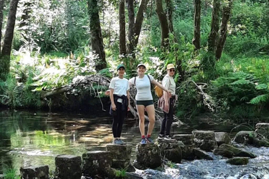 Tres mulleres de pé sobre as pedras pasadoiras dun río pouco fondo nunha fraga verde e soleada