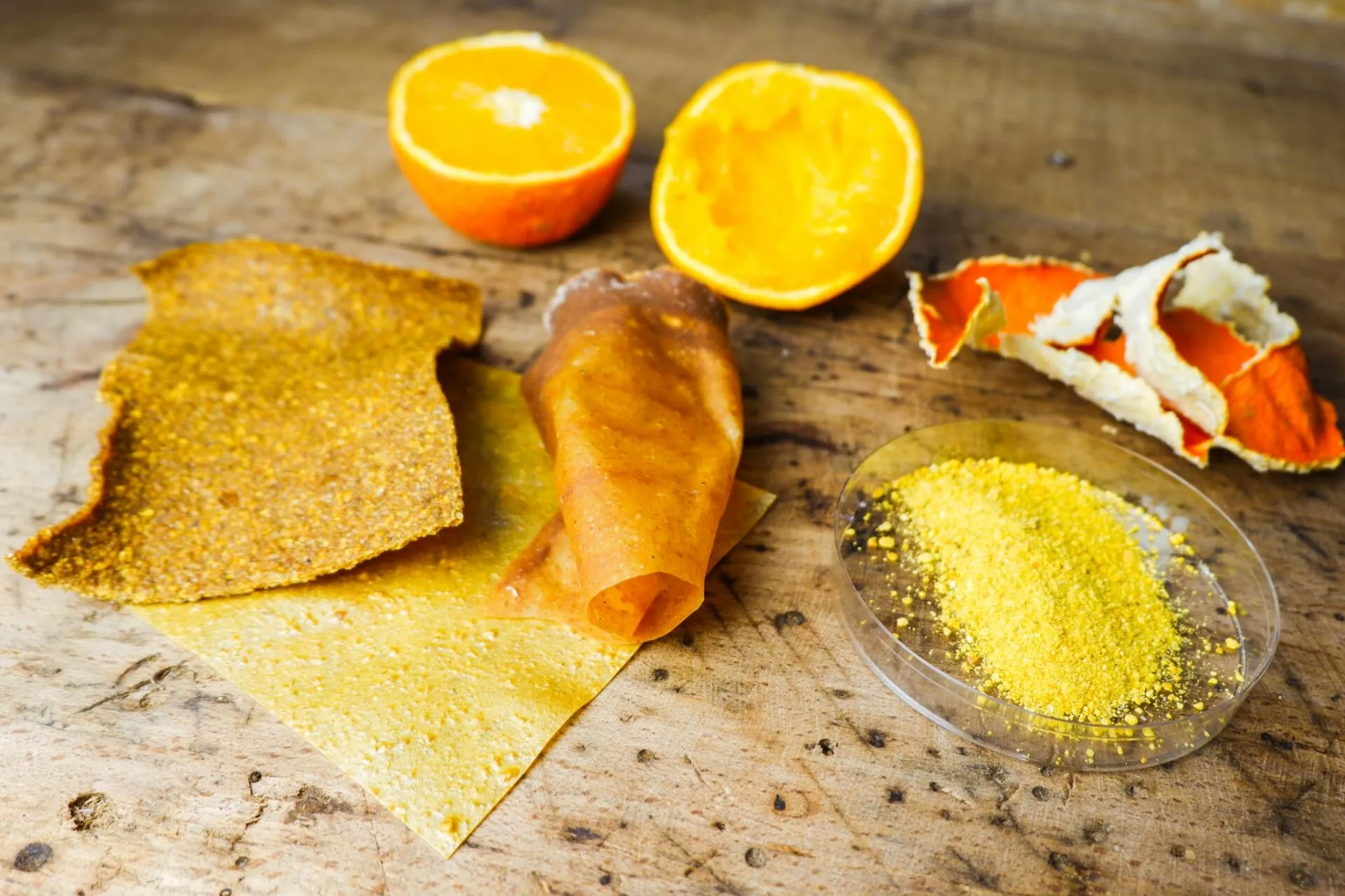 Naranjas partidas por la mitad, piel de naranja y láminas de fruta deshidratada sobre una mesa de madera rugosa, con un plato de polvo amarillo