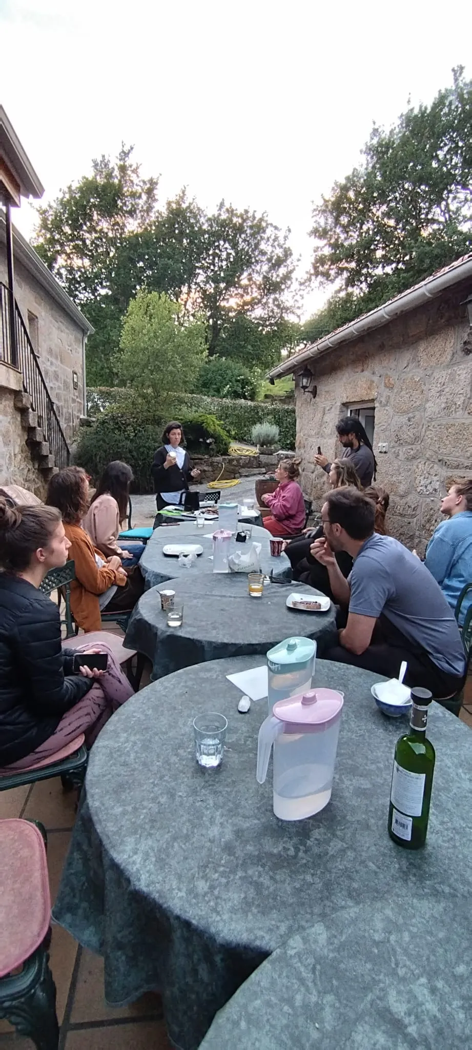 Una mujer habla a un grupo sentado alrededor de mesas redondas en una terraza de piedra al atardecer