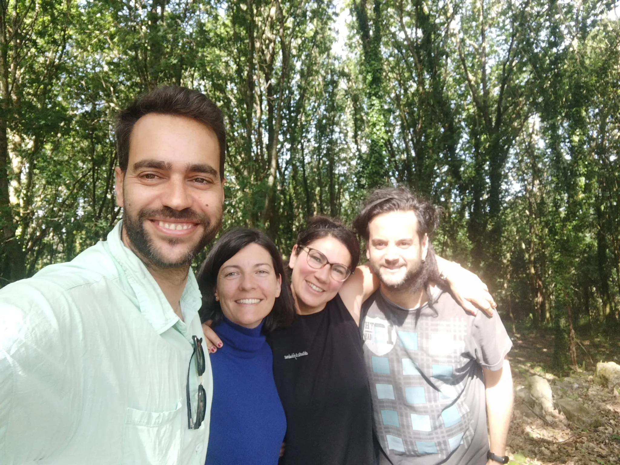 Selfi de cuatro personas sonrientes abrazadas en un bosque verde y soleado