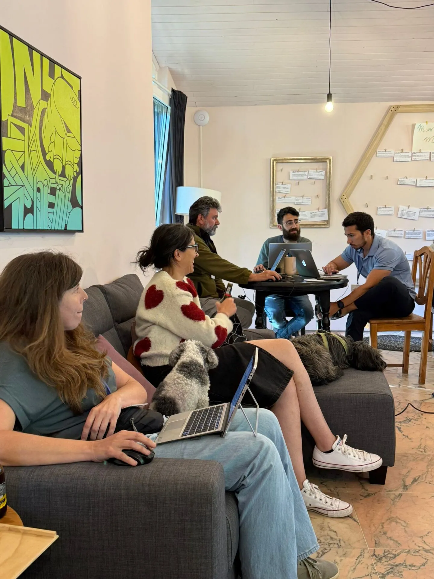 Seis persoas traballando cos portátiles en sofás e nunha mesa redonda, con dous cans descansando ao seu carón