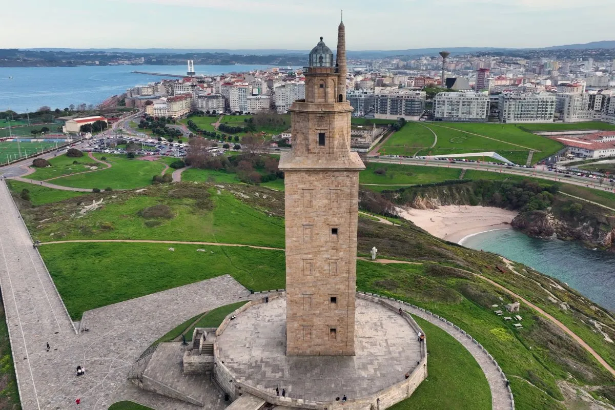 Vista aérea da Torre de Hércules sobre o seu promontorio verde, coas praias e o perfil urbano da Coruña ao fondo