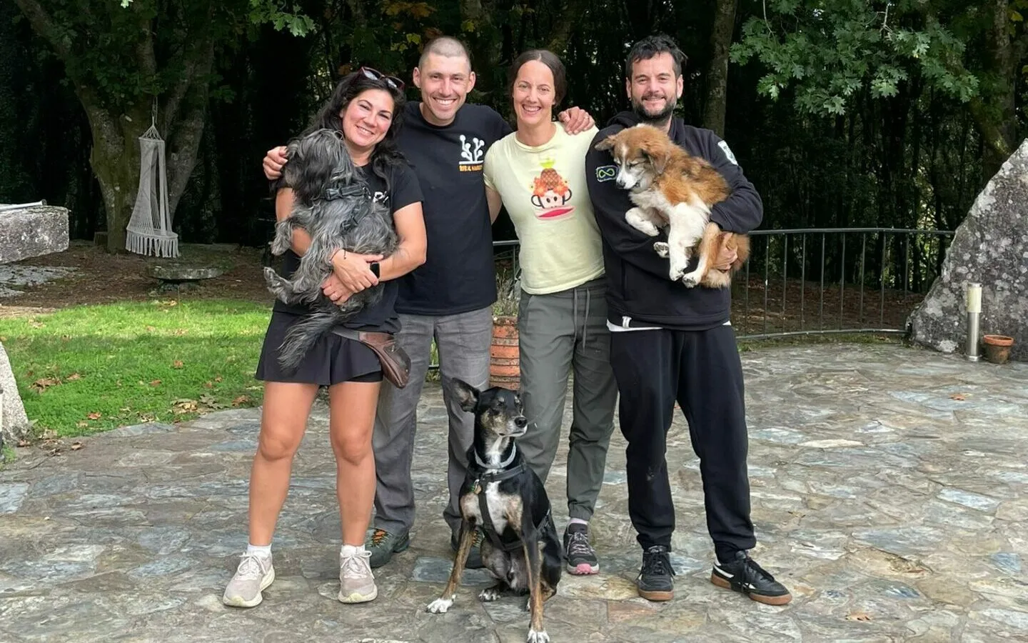 Catro persoas de pé abrazadas nunha terraza de pedra, dúas con cans pequenos no colo e un terceiro can sentado diante