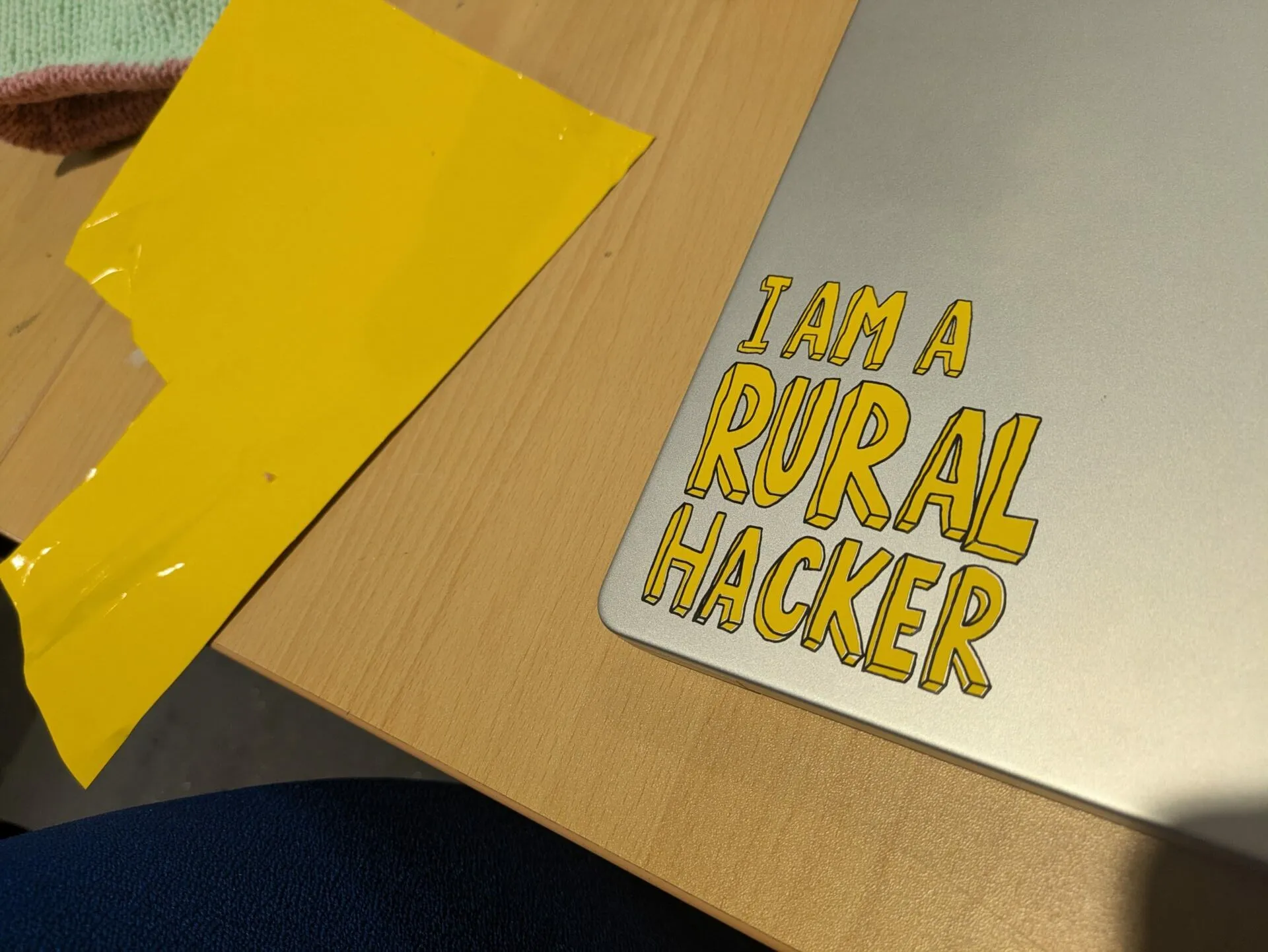 La tapa de un portátil con una pegatina amarilla que dice I AM A RURAL HACKER, sobre una mesa de madera junto a un trozo de cinta amarilla