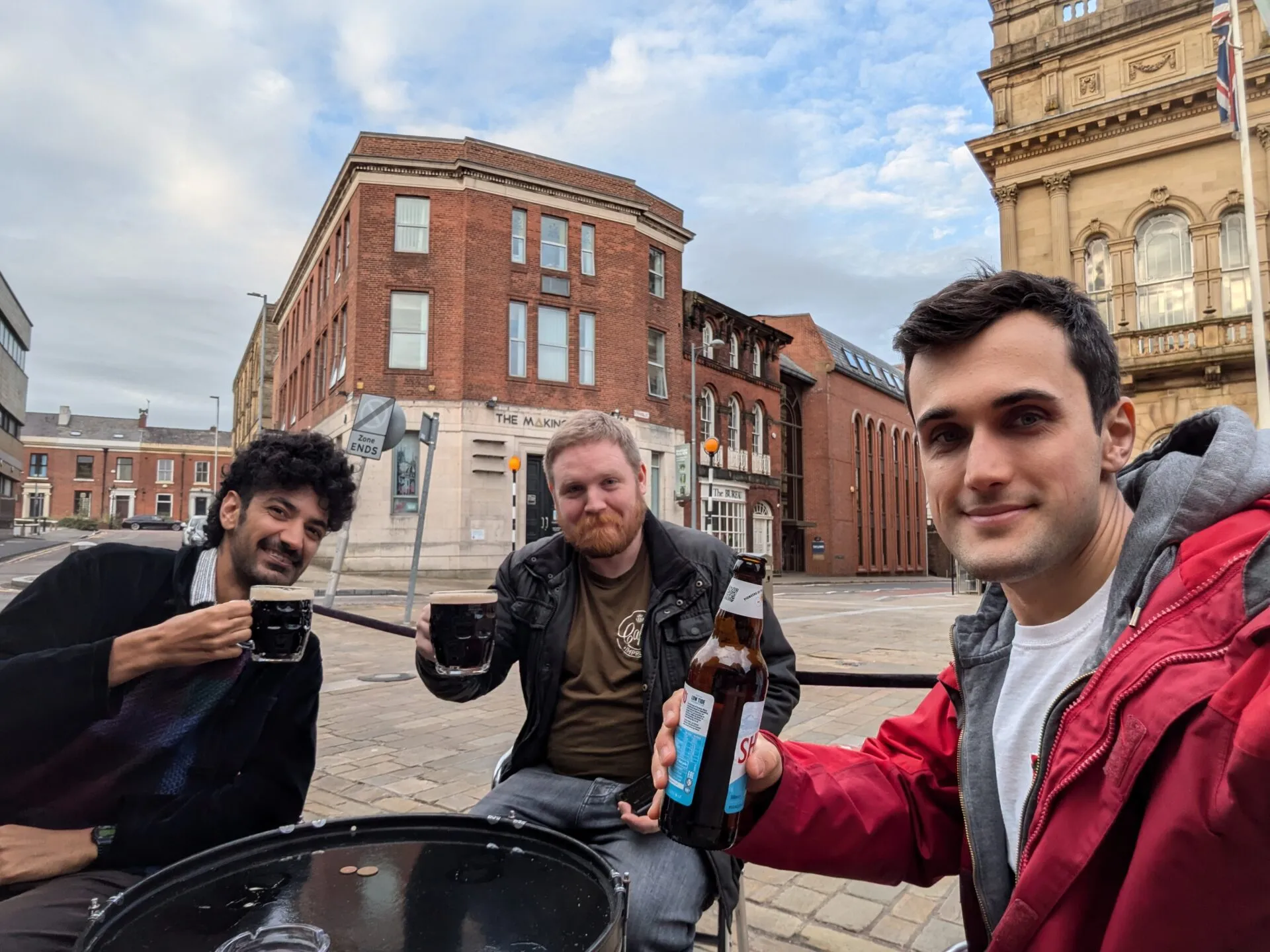 Tres hombres brindando con cervezas en una mesa exterior de una calle británica, con edificios de ladrillo rojo y una bandera del Reino Unido detrás