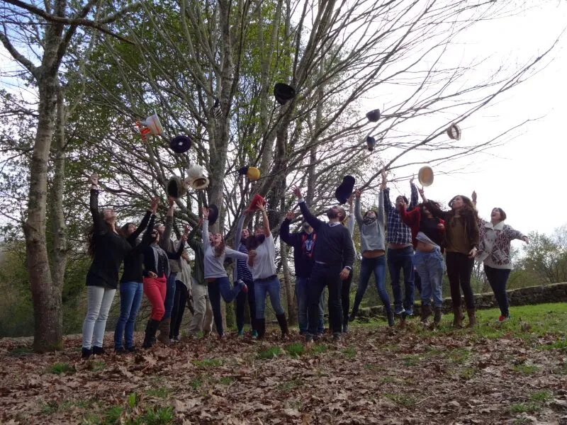 Unha quincena de persoas lanzando sombreiros ao aire baixo árbores sen follas, nunha ladeira cuberta de follas secas