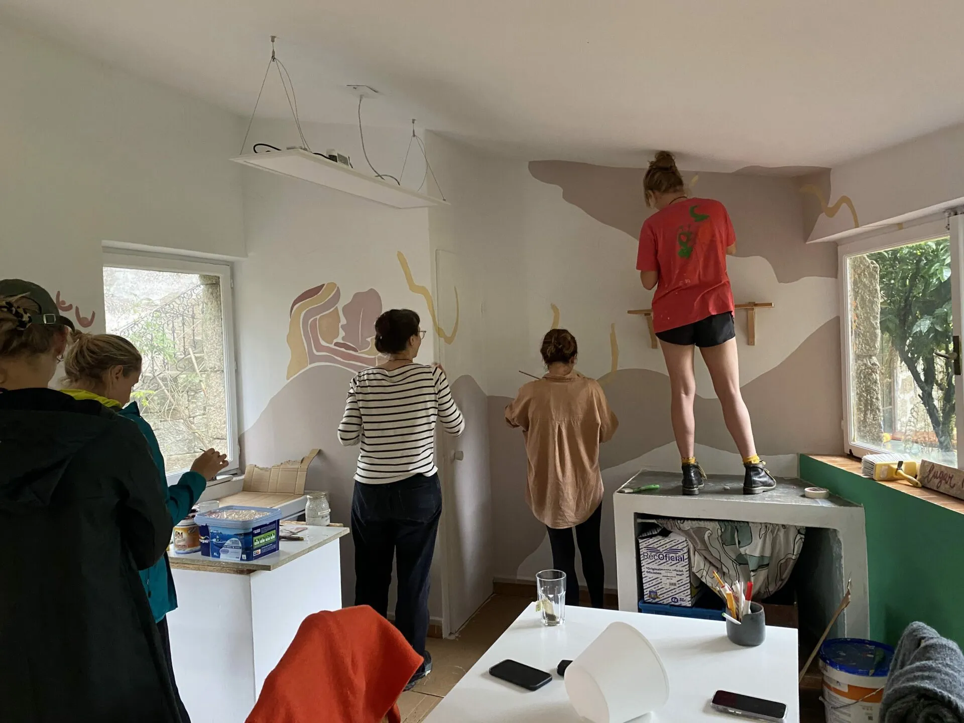 Catro persoas pintando as paredes do atelier en rosa pálido e ocre; unha está de pé na bancada para chegar ao teito