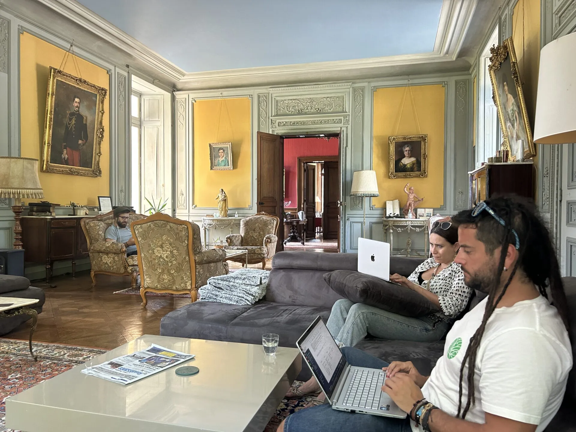 Tres persoas traballando cos portátiles no salón dun château, con paredes amarelas e retratos en marcos dourados