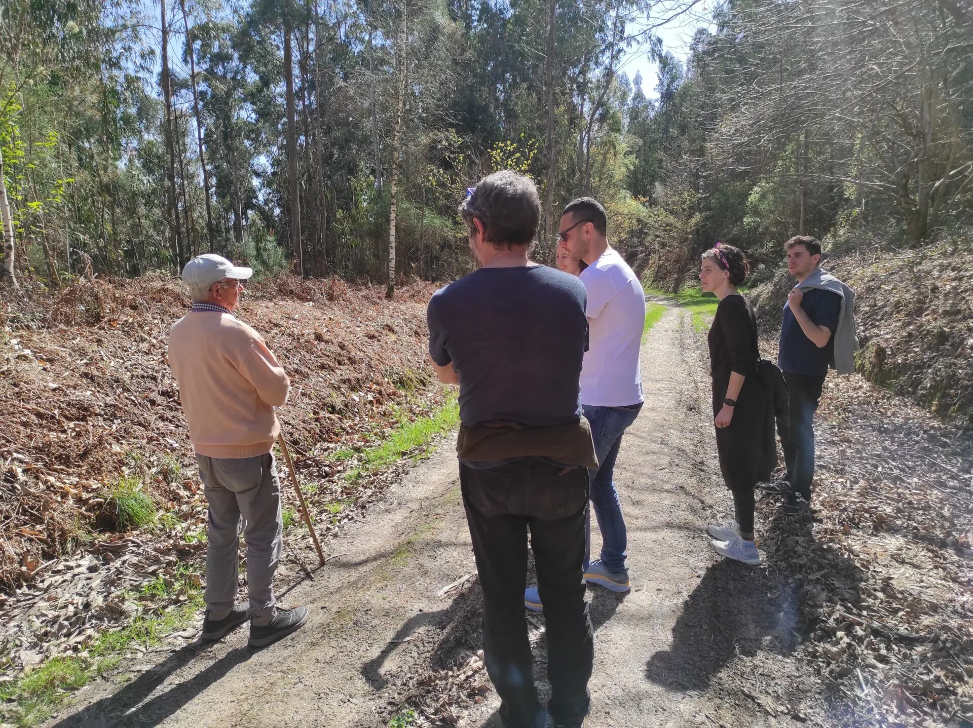 Hombre mayor con gorra blanca apoyado en un bastón hablando con cinco visitantes en una pista forestal soleada