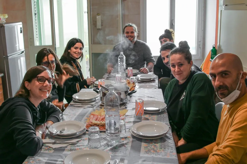 Nueve personas alrededor de una mesa larga en una cocina luminosa, con el vapor saliendo de una olla en el centro