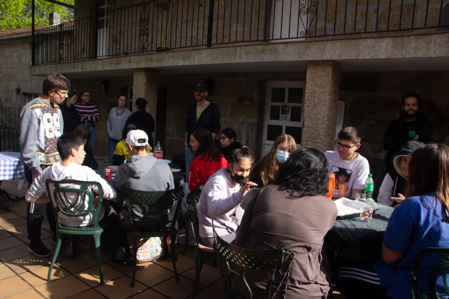 Mozos e adultos sentados en mesas nun patio de pedra soleado baixo un balcón, algúns con máscara