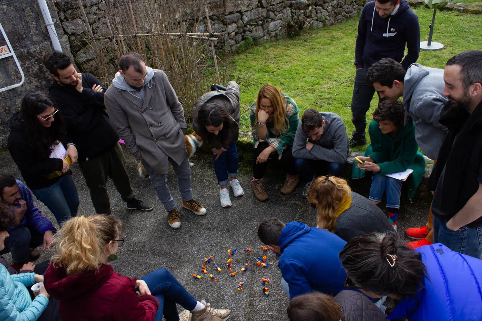 Vista desde arriba, un gran círculo de personas de pie y en cuclillas alrededor de pequeñas construcciones de Lego en un patio de cemento