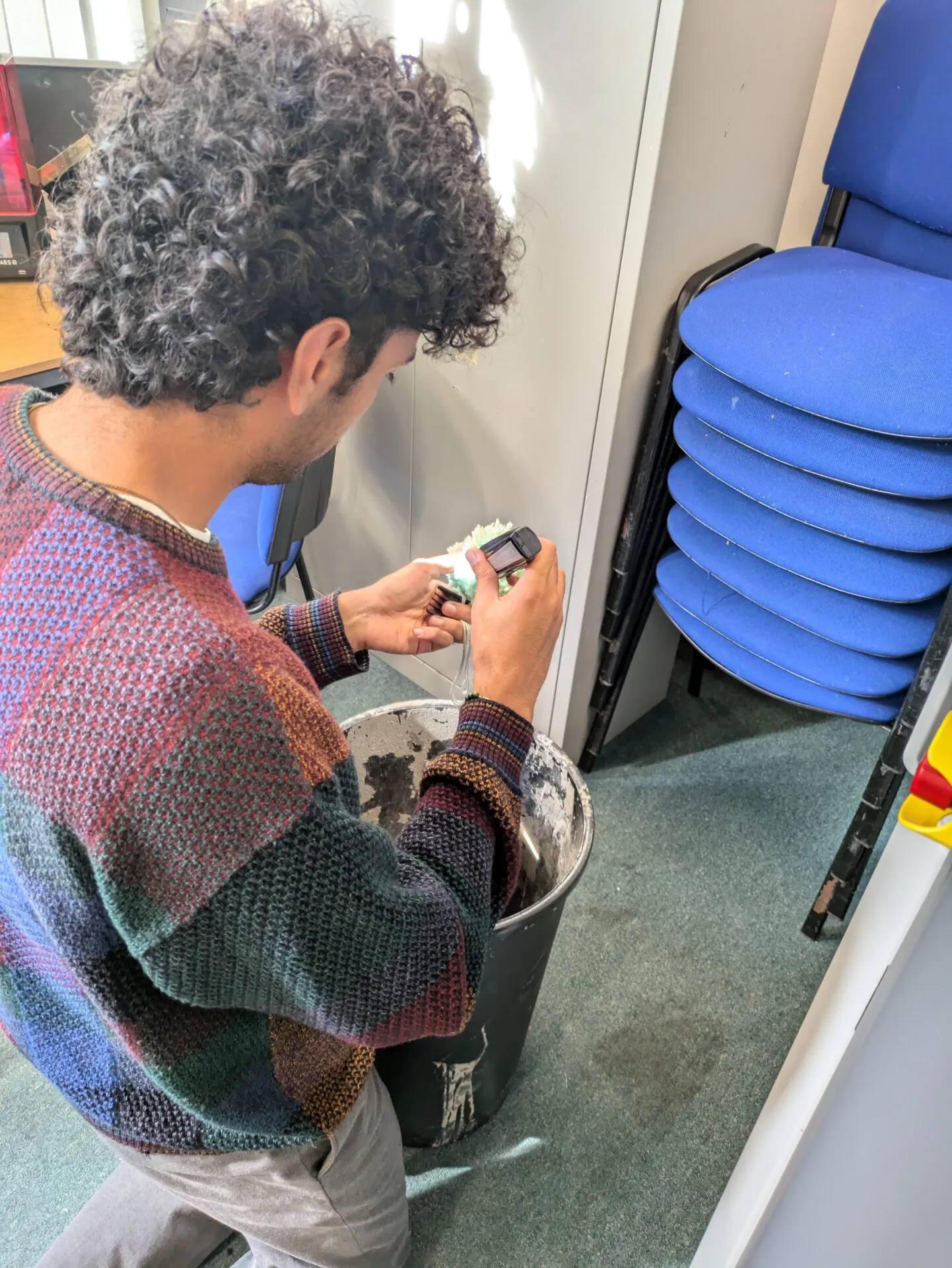 Hombre con jersey de cuadros examina un pequeño aparato y una pieza de plástico moteada de verde sobre un cubo en un taller