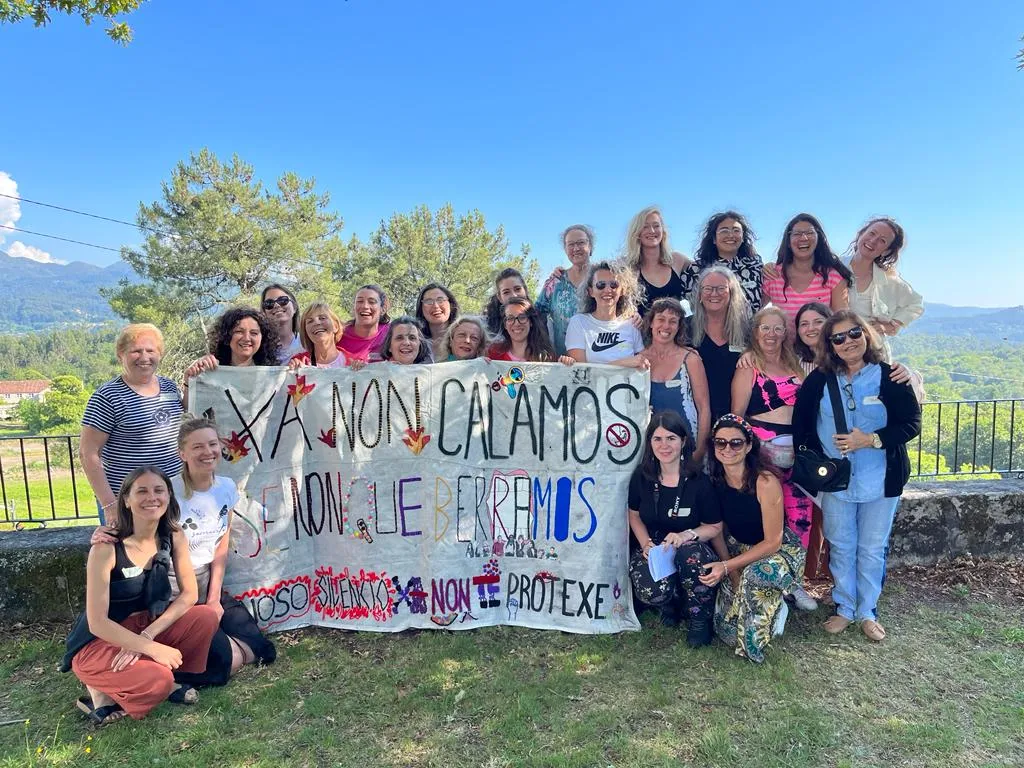 Un grupo de mulleres posando nun céspede tras unha pancarta pintada a man que di Xa non calamos, cunha cerca e montes detrás