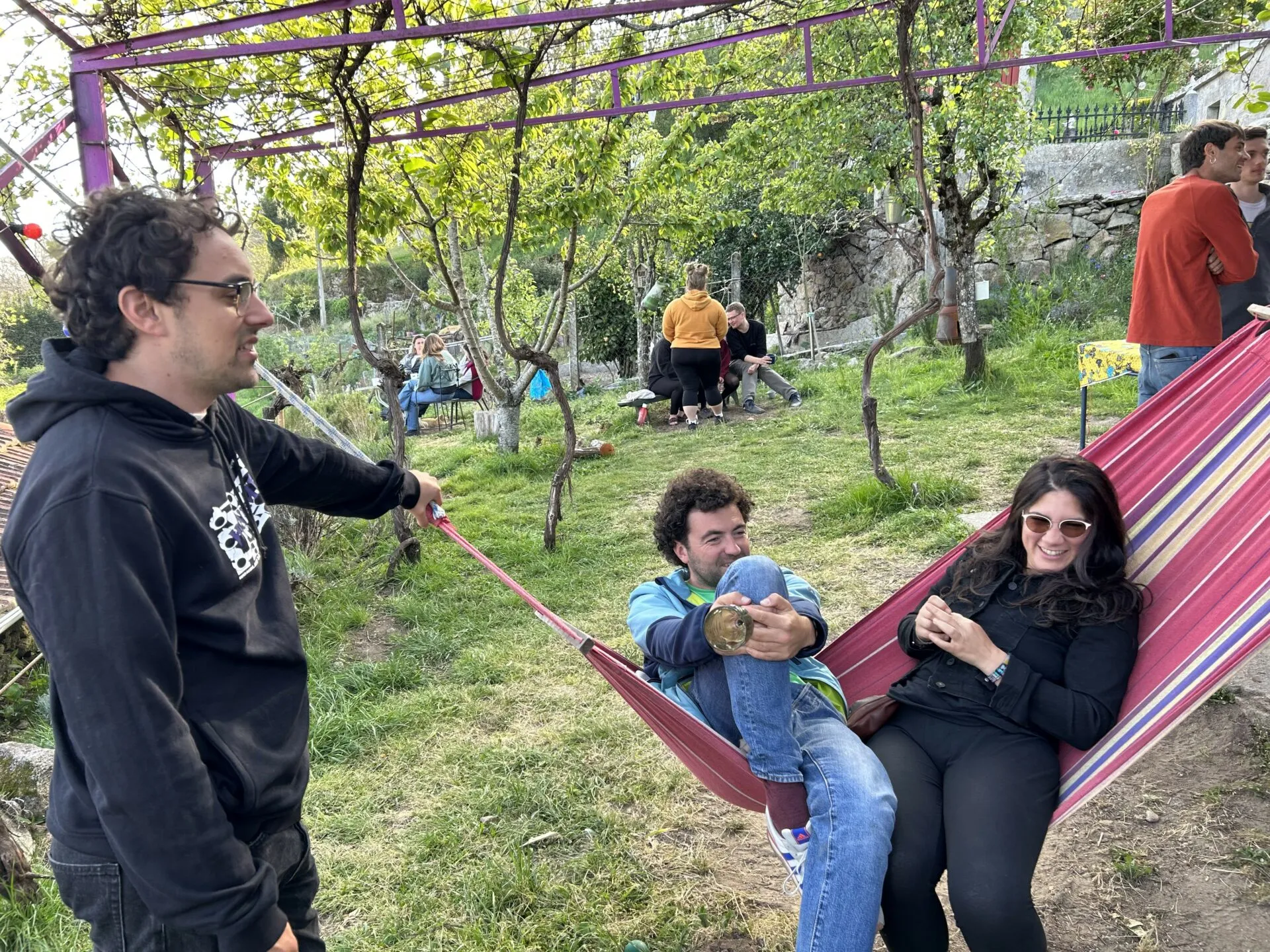 Dos personas sentadas en una hamaca de rayas bajo una pérgola en un jardín soleado mientras un hombre con sudadera sujeta la hamaca