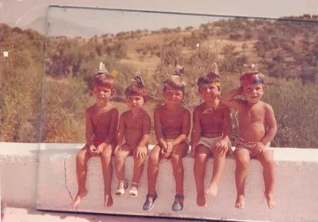 Fotografía antiga descolorida de cinco nenos pequenos en traxe de baño sentados en fila sobre un muro branco baixo, con outeiros secos detrás