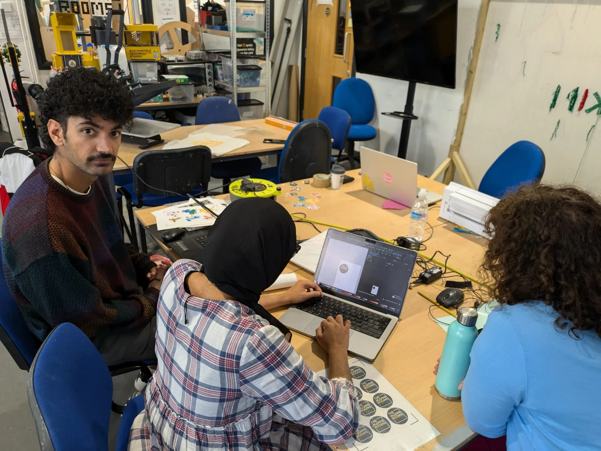 Tres personas trabajando con portátiles y hojas de pegatinas en una mesa abarrotada de un makerspace