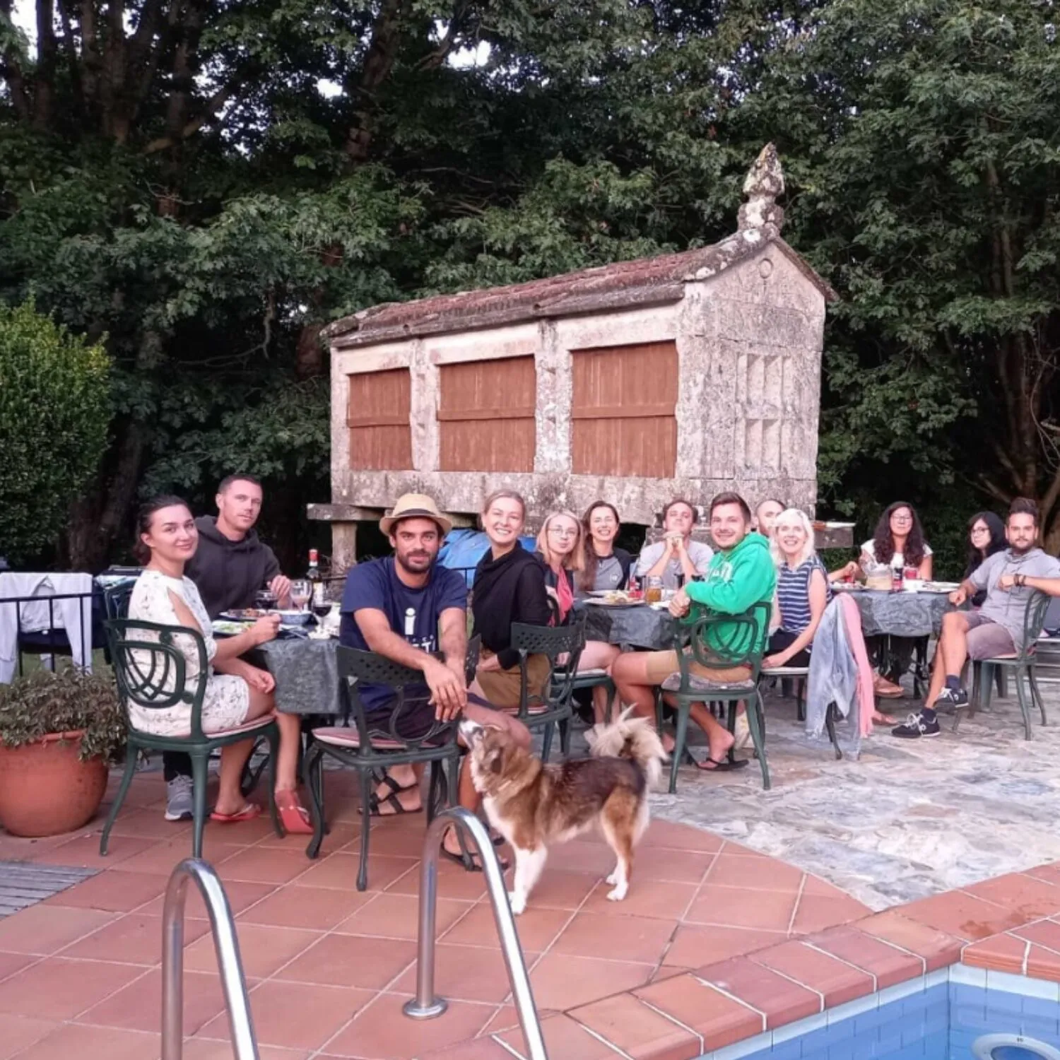 Unhas quince persoas ceando en mesas ao aire libre a carón dunha piscina, diante dun hórreo de pedra