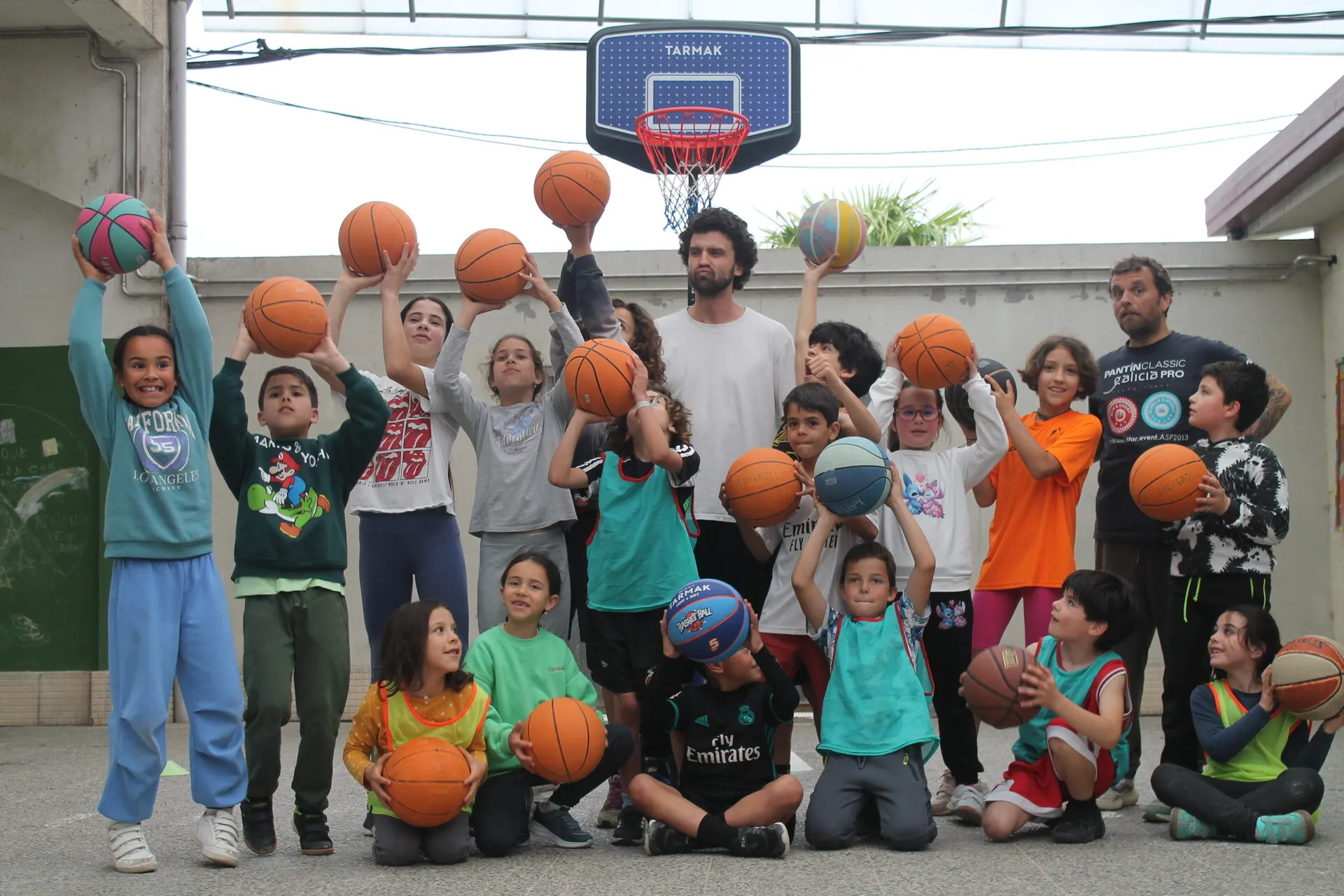 Una veintena de niños levantando balones de baloncesto bajo una canasta en un patio de colegio, con Víctor y otro entrenador detrás