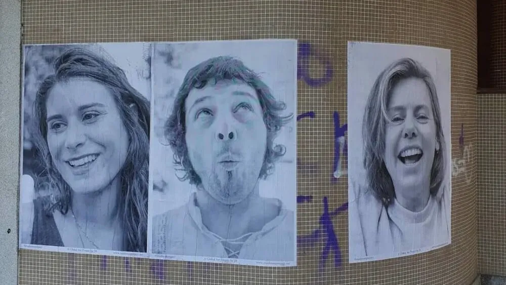 Tres grandes carteis en branco e negro con caras rindo pegados nunha parede de azulexos cuberta de grafitis morados