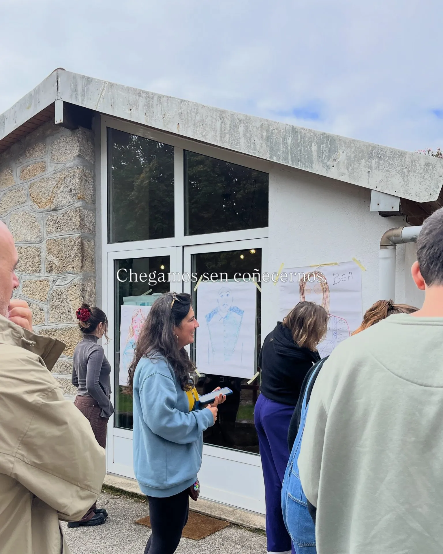 People gathered outside a small building of stone and white walls, looking at drawn portraits taped to its glass door and window; a woman in a light blue sweatshirt smiles as she holds a phone