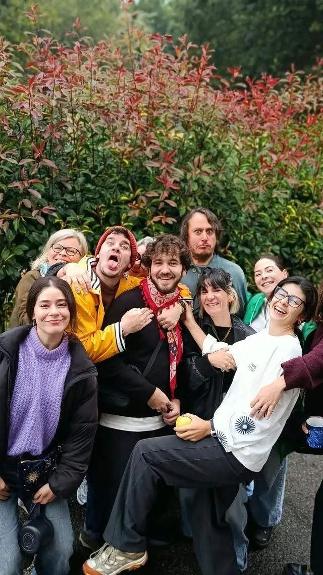 A large group of people posing outdoors in front of a hedge with red and green leaves, laughing and hugging; a man in a red beanie and yellow jacket sticks out his tongue