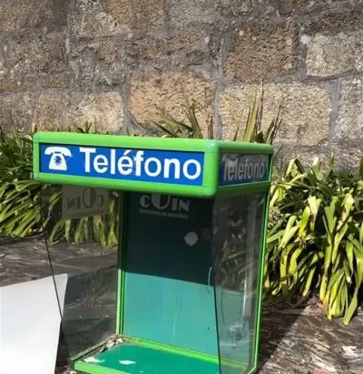 An old green public phone booth marked “Teléfono” standing on stone paving in front of a granite wall and green plants