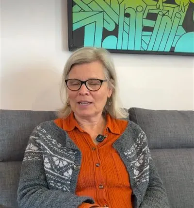 An older woman with fair hair and glasses, in a patterned grey cardigan over an orange shirt, sitting on a grey sofa under a green and blue painting and speaking to the camera