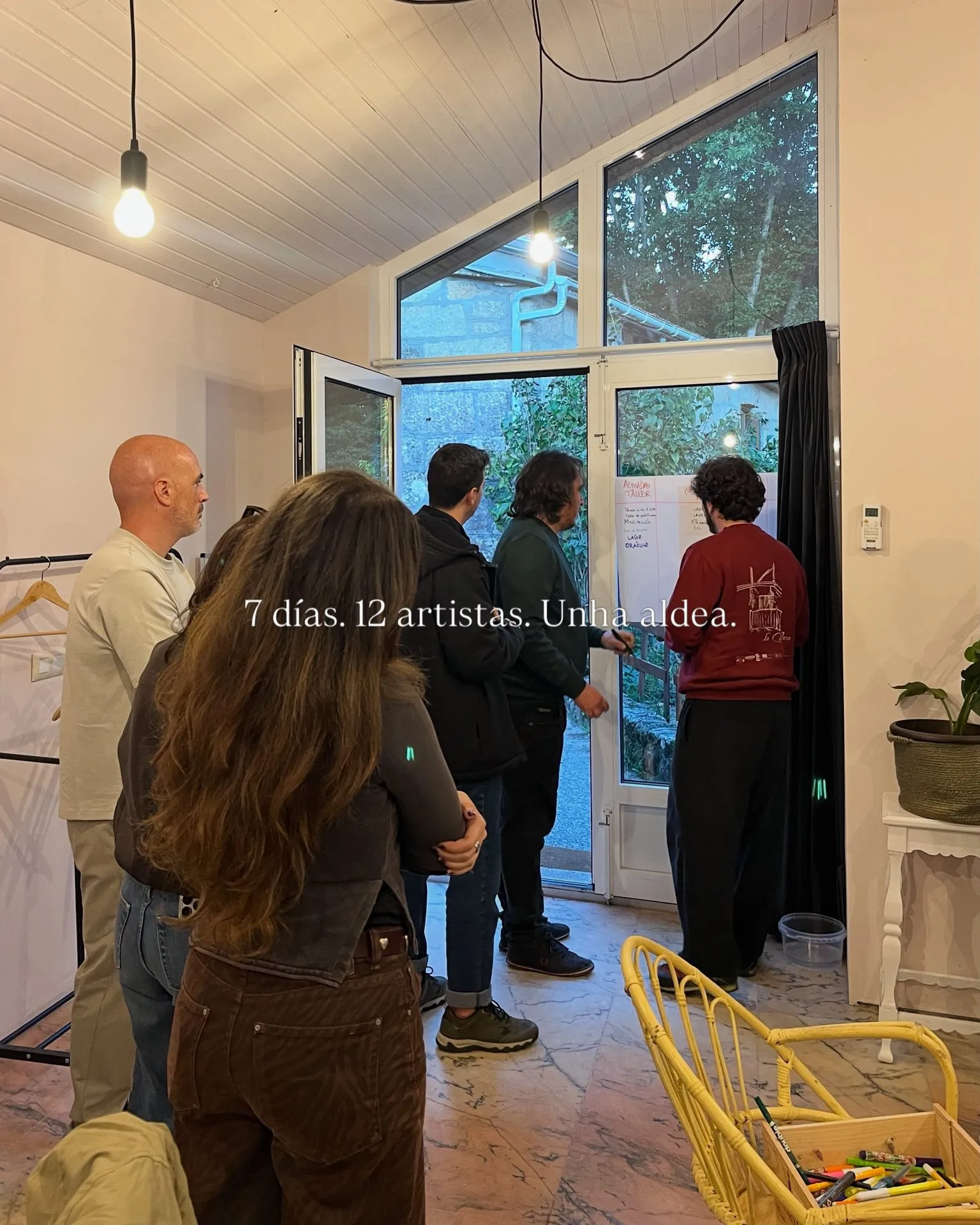 A group of people standing in a bright room with large windows and hanging bulbs, watching a man write on a sheet of paper stuck to the glass door; overlaid text reads “7 días. 12 artistas. Unha aldea.”